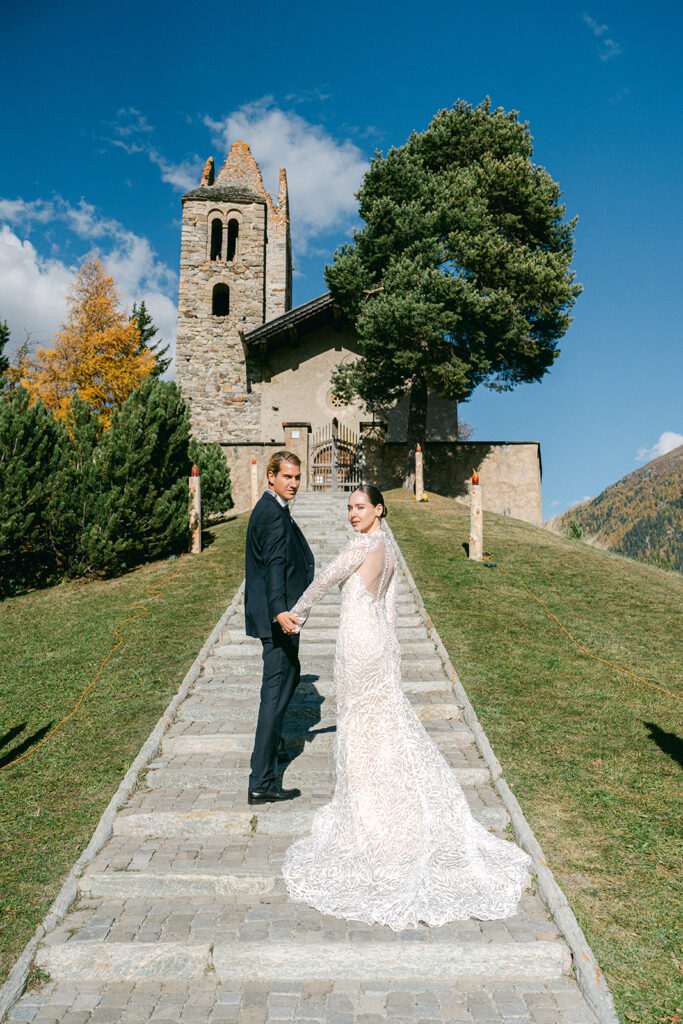 Eine Braut und ein Bräutigam halten sich auf den Steinstufen vor einer rustikalen Kirche in der Schweiz an den Händen, umgeben von Bäumen und Hügeln unter einem blauen Himmel - perfekt für eine Hochzeitssängerin oder Traurednerin, um ihrem besonderen Tag einen besonderen Zauber zu verleihen.
