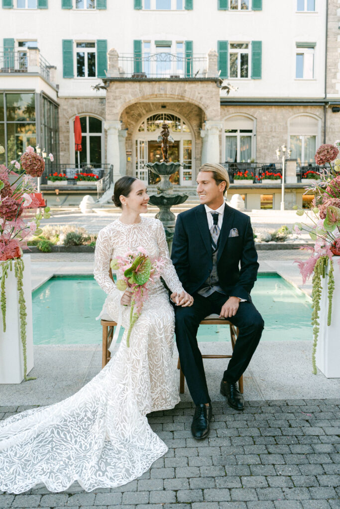 Die Braut in einem weißen Kleid und der Bräutigam in einem schwarzen Anzug sitzen lächelnd an einem Brunnen und halten sich an den Händen, während eine Hochzeitssängerin auf ihrer Schweizer Hochzeit auftritt.