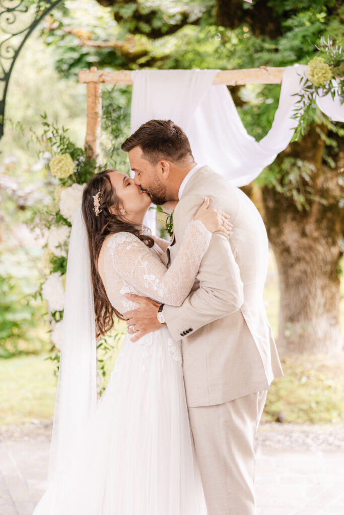 The bride and groom kiss during their outdoor wedding ceremony under a floral arch, while a wedding singer from Zurich serenades them.