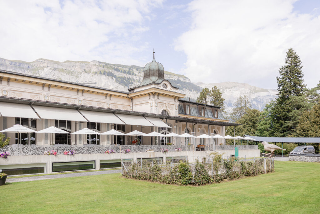 Historic building with a dome against the backdrop of the Swiss Alps, with umbrellas on the terrace and a green meadow in front.