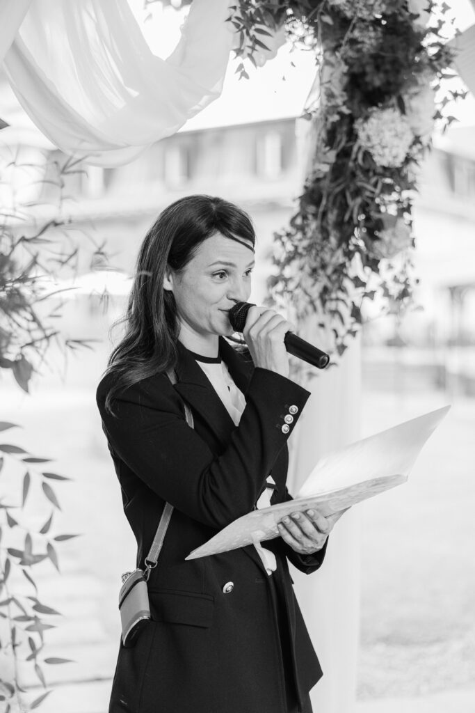 A female officiant in a suit speaks into a microphone while holding papers at an outdoor event under a flower arch in Switzerland.