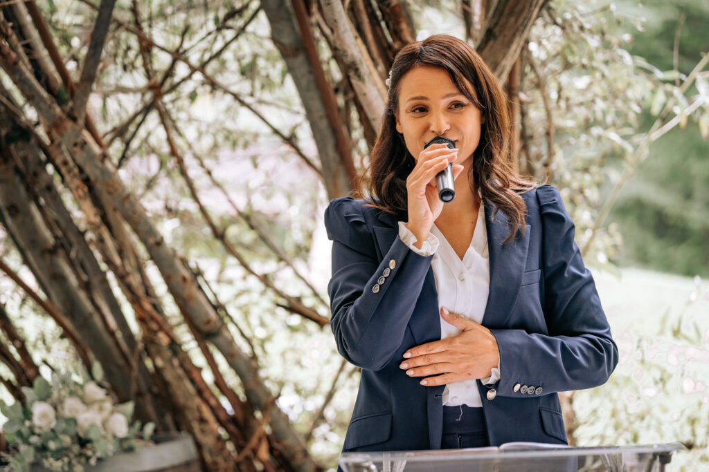 Naomi Ben speaks as a wedding DJ with a microphone in a green setting, surrounded by trees - an atmospheric ceremony in Zurich.