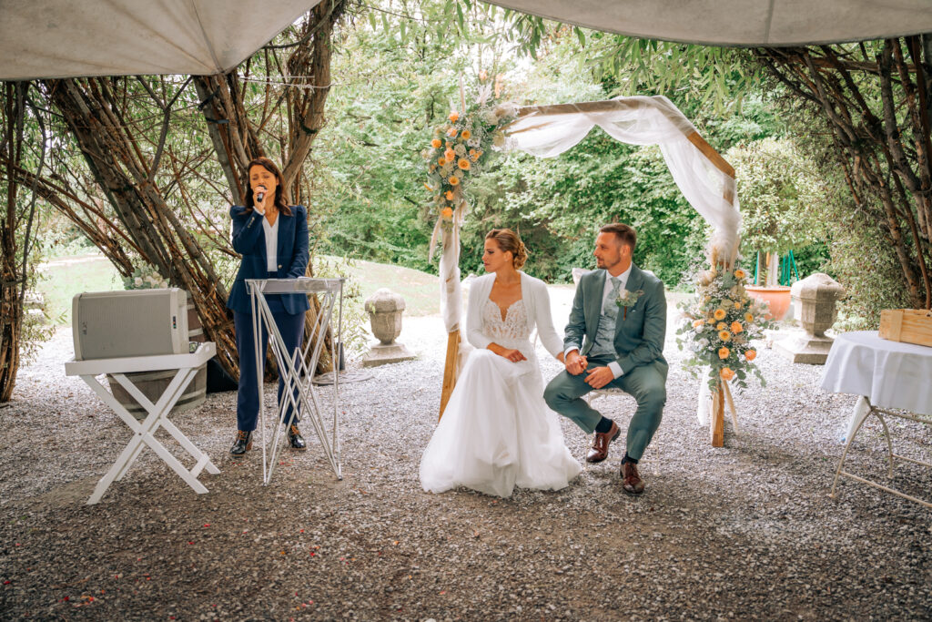 The bride and groom are sitting under a floral arch in Switzerland, while a celebrant speaks during their outdoor wedding ceremony.