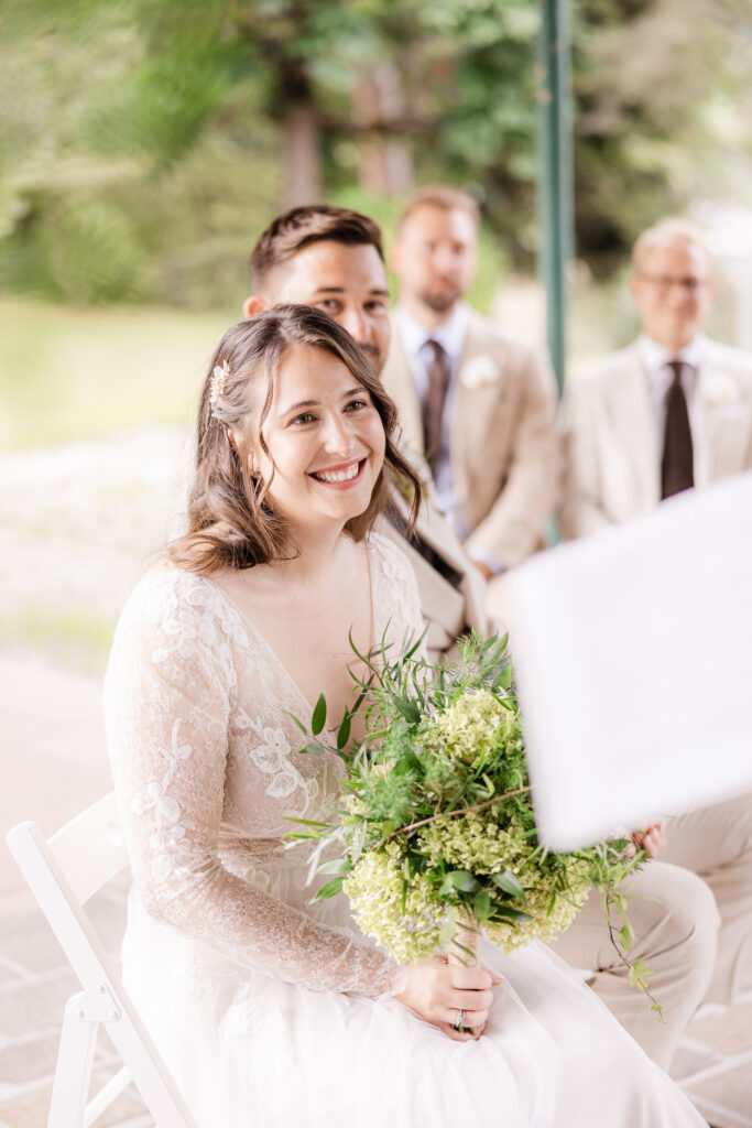Eine lächelnde Braut im Spitzenkleid hält einen Blumenstrauß, während die Trauzeugen im Hintergrund auf einer Hochzeit in Zürich, Schweiz, sitzen - bereit, mit dem perfekten Hochzeits-DJ zu feiern.