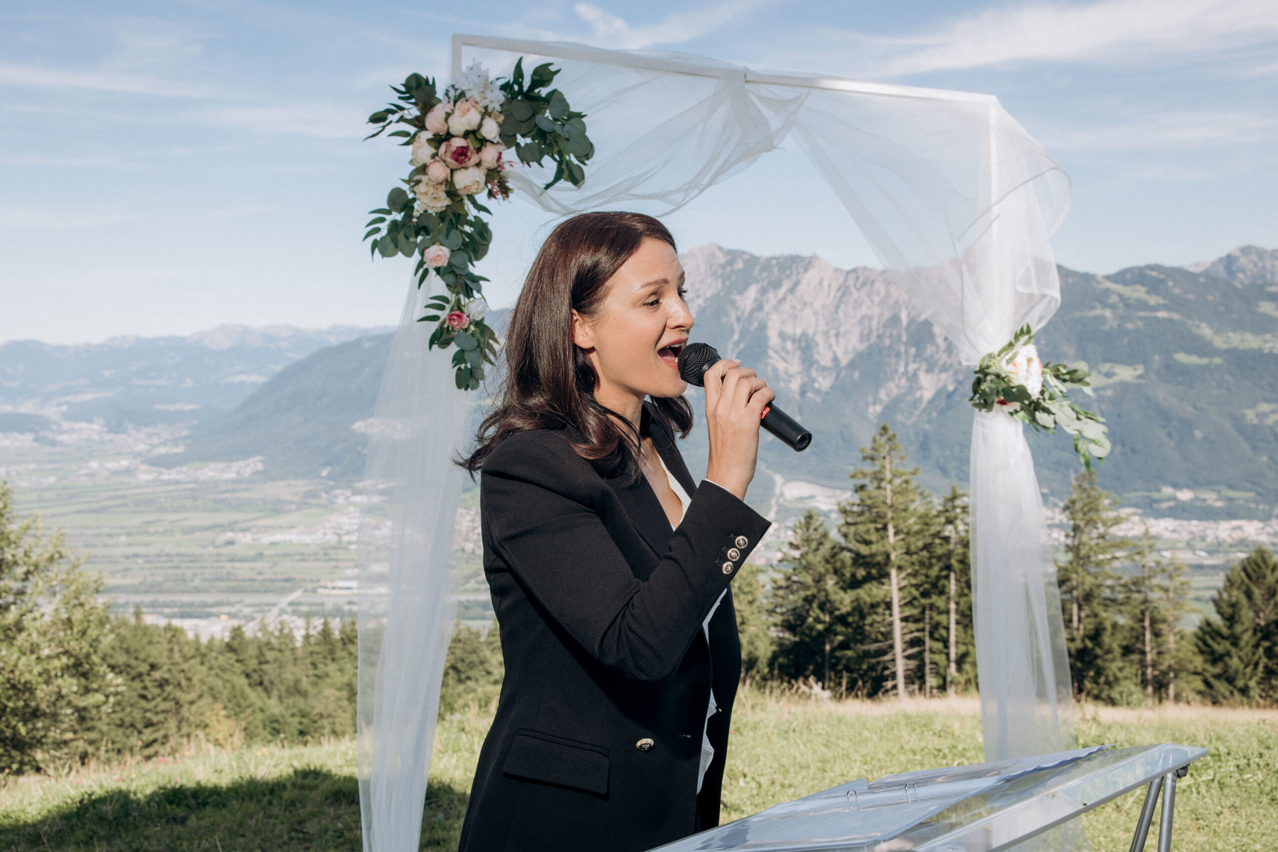 Female officiant in a black blazer speaking into a microphone outdoors, with a breathtaking mountain backdrop and a wedding arch in Switzerland.