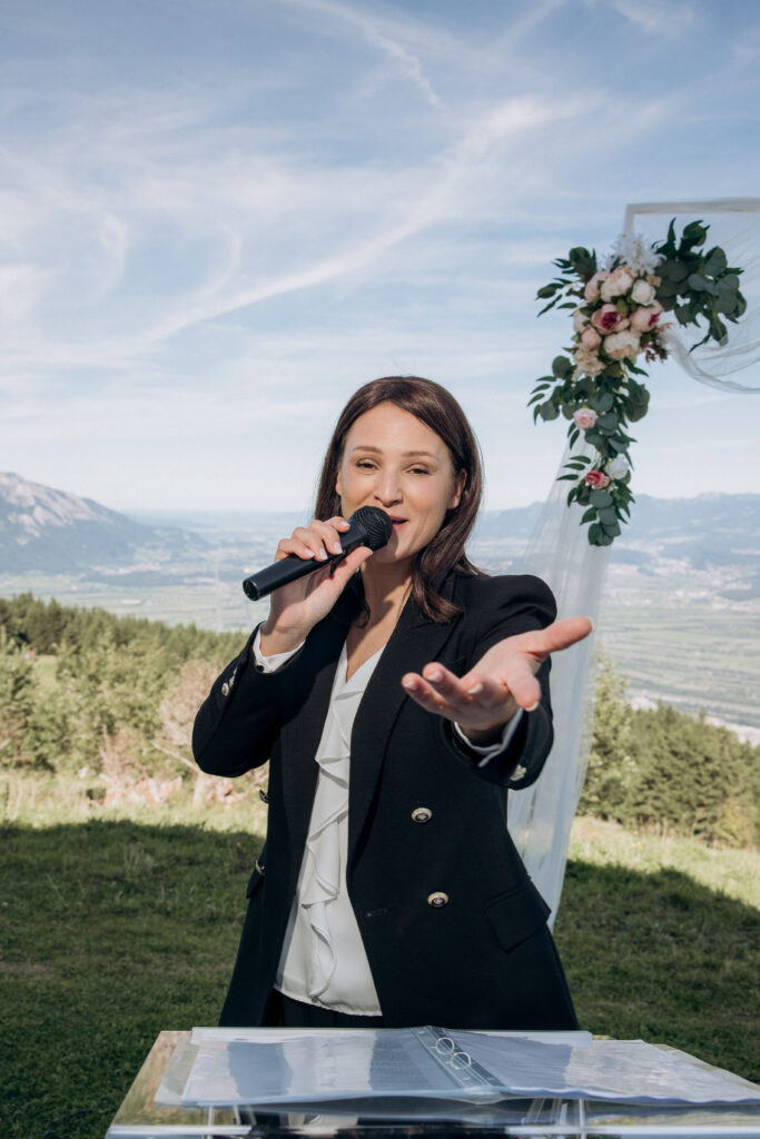 A wedding singer in an elegant suit speaks into a microphone, gesturing in front of a floral wedding arch as she performs at an outdoor ceremony in Zurich.