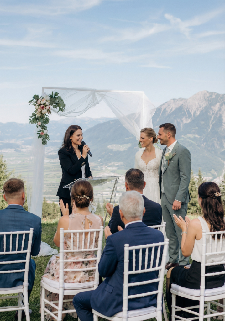 A couple stands under a floral arch during an outdoor wedding ceremony in Zurich, as guests watch and applaud, accompanied by a talented wedding singer.
