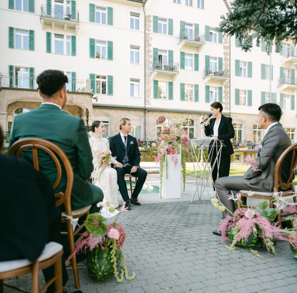 Outdoor wedding in Zurich with a seated couple, a wedding officiant performing the ceremony, and guests watching in a charming courtyard.